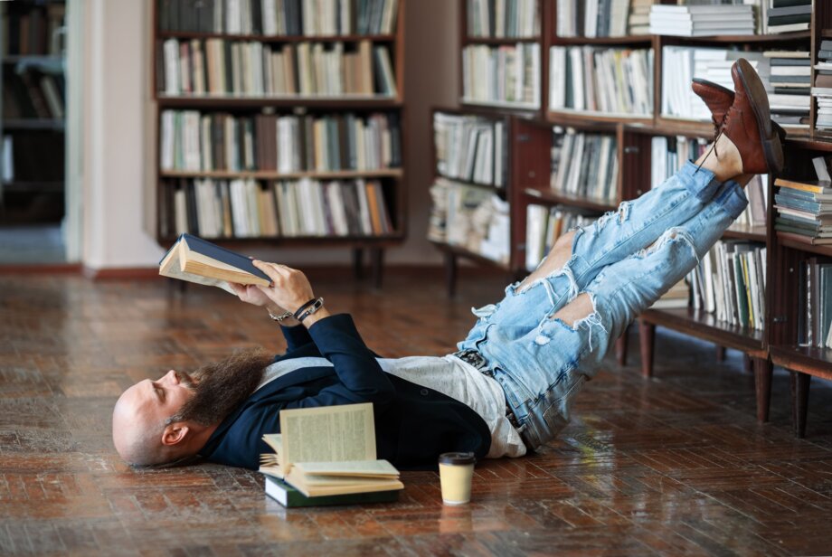 Mężczyzna leżący na podłodze z nogami założonymi na biblioteczce. W rękach trzyma książkę, którą czyta