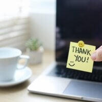 Dłoń trzymająca karteczkę z napisem "thank you" na tle laptopa